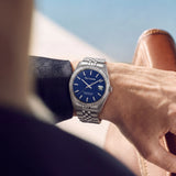 A lifestyle photograph of the Guy Laroche Rosalie stainless steel watch on a man's wrist, pairing with a tailored dark navy jacket and leather loafers to highlight the vibrant blue dial and fluted bezel.