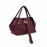 Left three-quarter studio view of a premium burgundy leather handbag featuring unique folded side wings, braided top handles, and a signature gold-tone logo emblem with a long leather tassel, available at BLUE SALON.