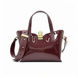 Front view of a burgundy patent leather handbag featuring a structured silhouette, dual top handles, a gold-tone clasp closure, and a wide matching shoulder strap slung across the front.