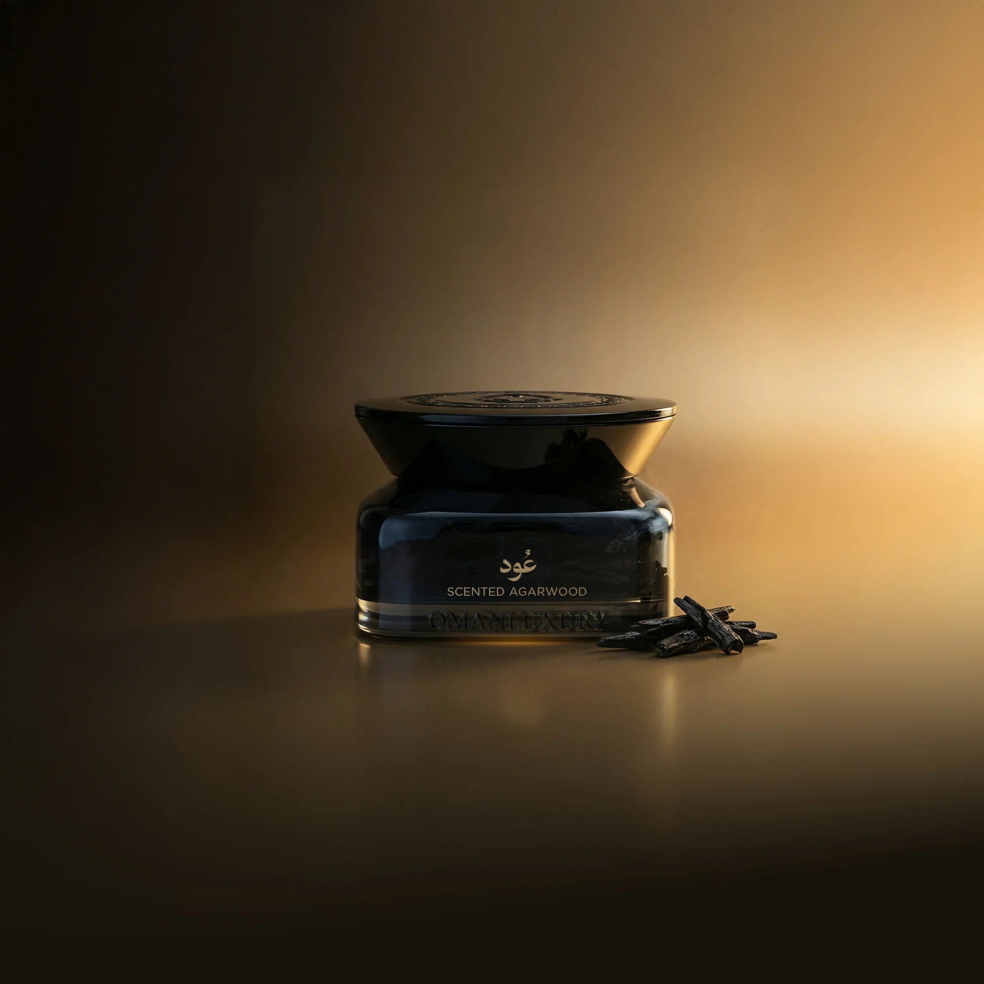 Dark glass jar with gold-toned ornate lid and 'عود SCENTED AGARWOOD' text. Dark, irregular agarwood pieces are next to it.