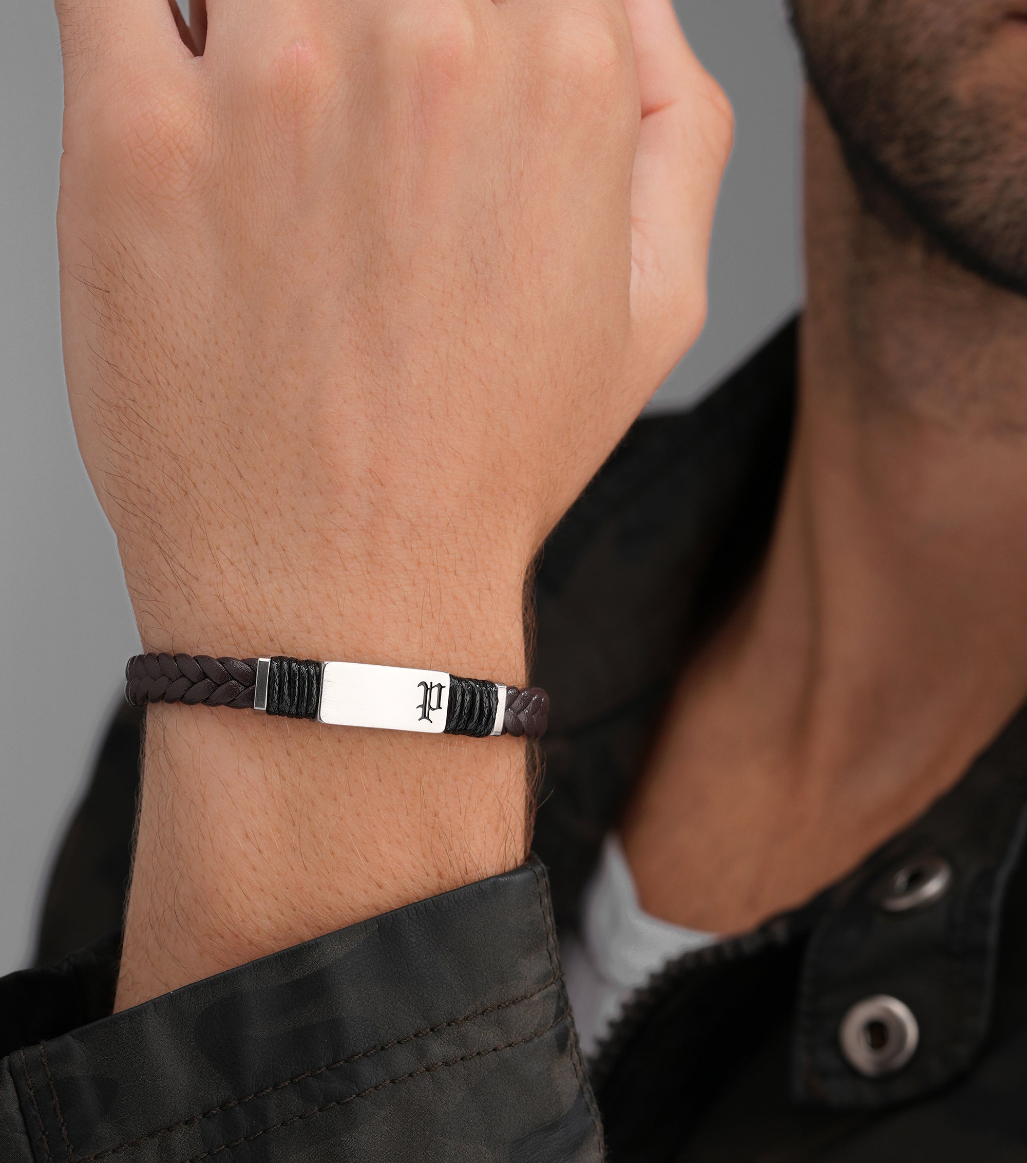 Dark brown braided leather bracelet with a silver-toned rectangular plate engraved with a gothic "P", flanked by black cord w