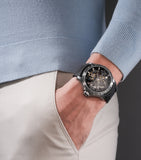 Gunmetal-toned stainless steel watch with a black skeleton dial showing silver-toned and gold-toned mechanisms, on a black si