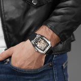 Silver-toned tonneau steel watch with a black skeleton dial showing gold-toned gears, on a black silicone strap.