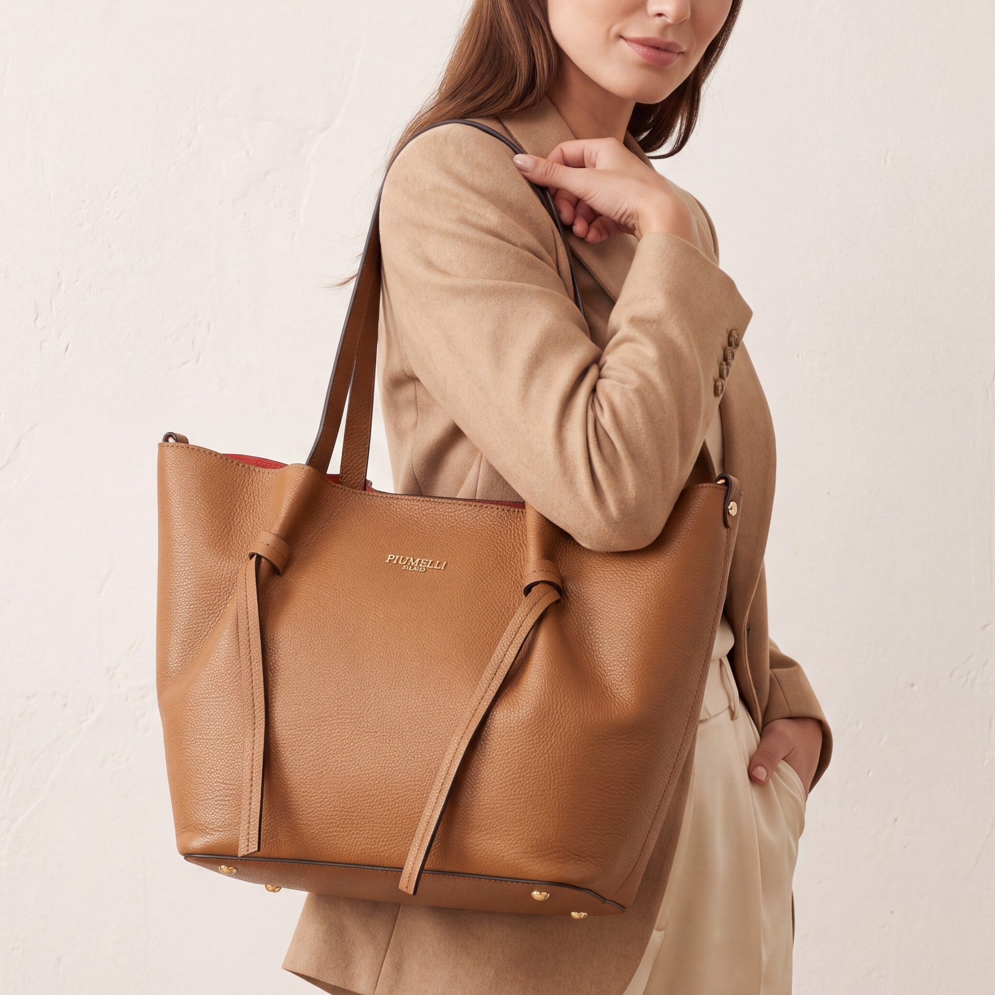 Warm brown pebbled leather tote bag with two shoulder straps, gold-toned logo, decorative front ties, and red lining.