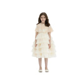 A young girl smiling in a champagne-colored, tea-length special occasion dress featuring a multi-layered tiered tulle skirt and a floral lace capelet with a ribbon tie, set against a white studio background.