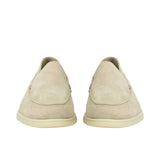 A straight-on, front-facing view of a pair of beige suede slip-on loafers, highlighting their rounded apron toe boxes and clean, symmetrical slip-on design resting on off-white rubber outsoles.