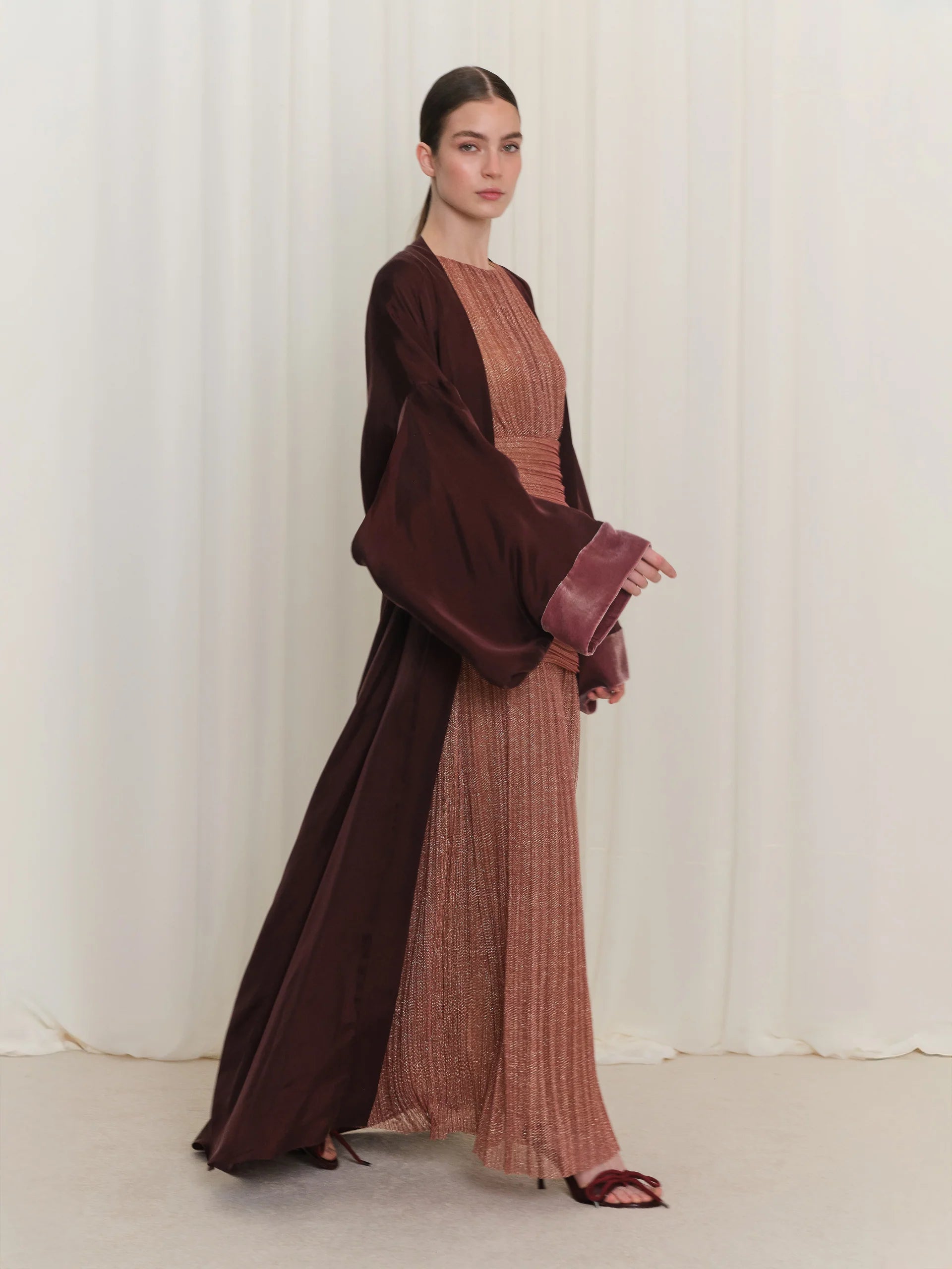 Burgundy open abaya with wide sleeves and lighter burgundy velvet-lined cuffs, over a rose gold pleated dress with a ruched b