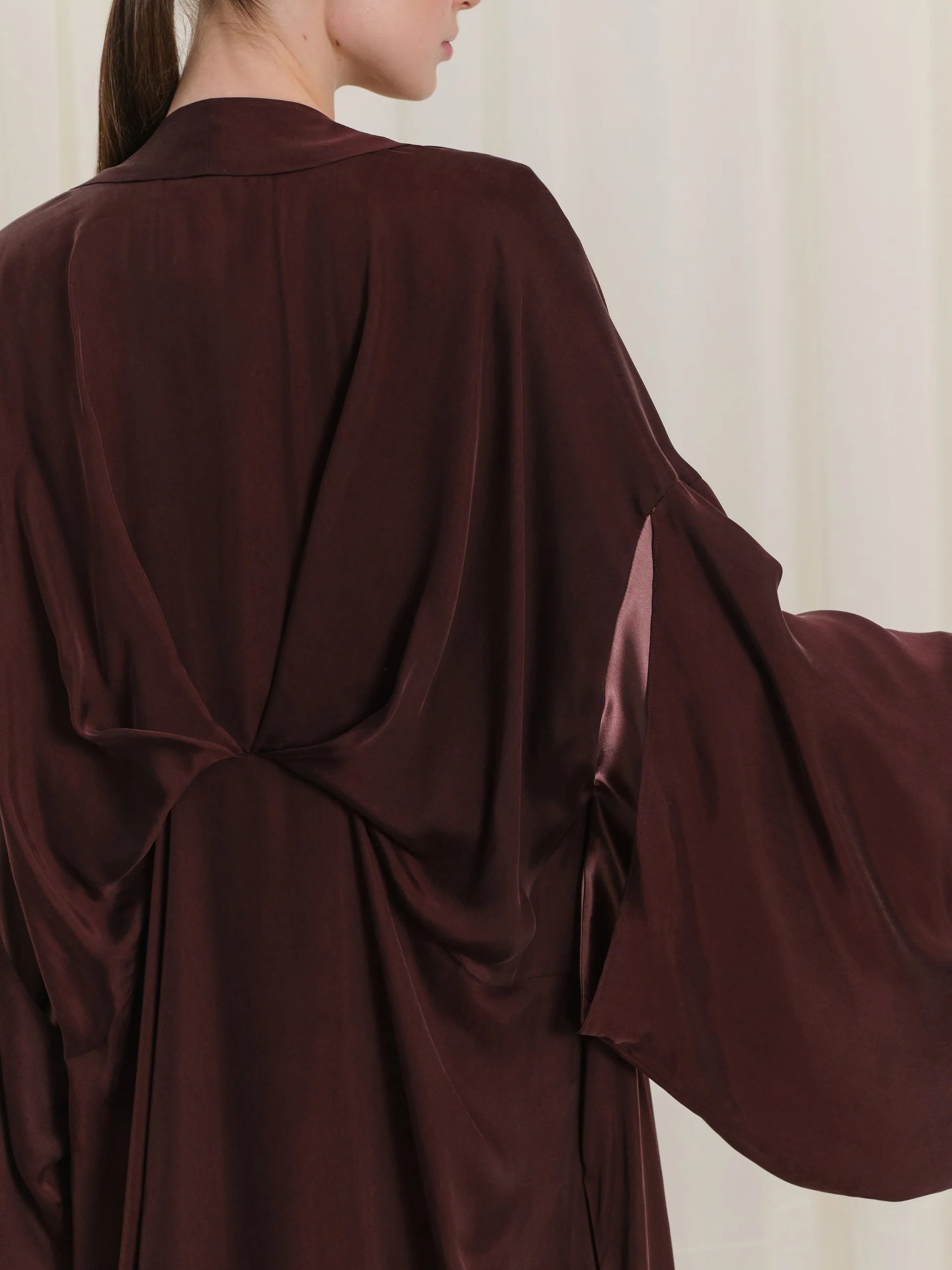 Burgundy abaya with a draped back, gathered at the center. Wide sleeves feature a slit revealing a lighter inner lining.