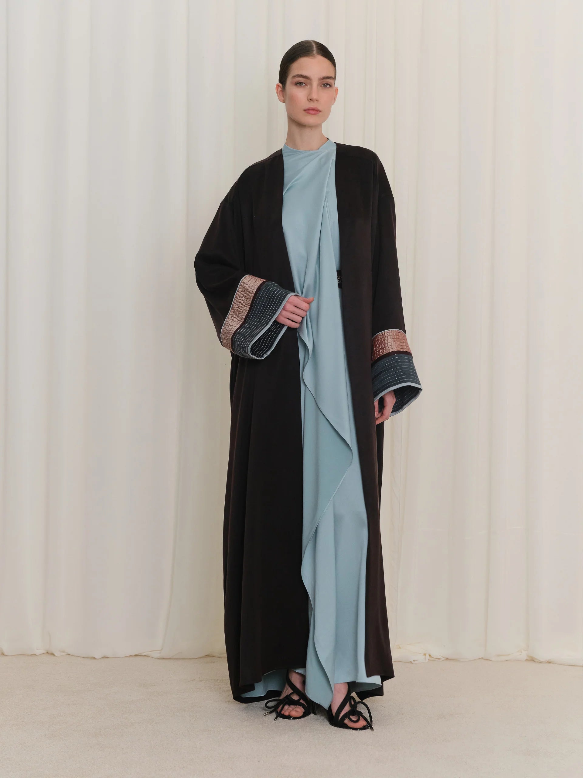 Black open abaya with long sleeves. Cuffs feature a rose gold-toned textured band and a dark blue striped section.