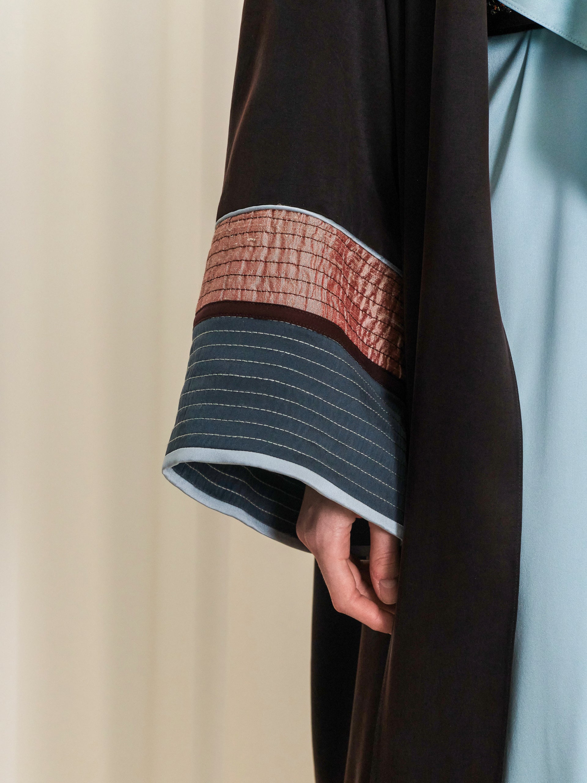 Black abaya sleeve with a crinkled reddish-brown section, a dark teal section with horizontal dashed stitching, and light blu
