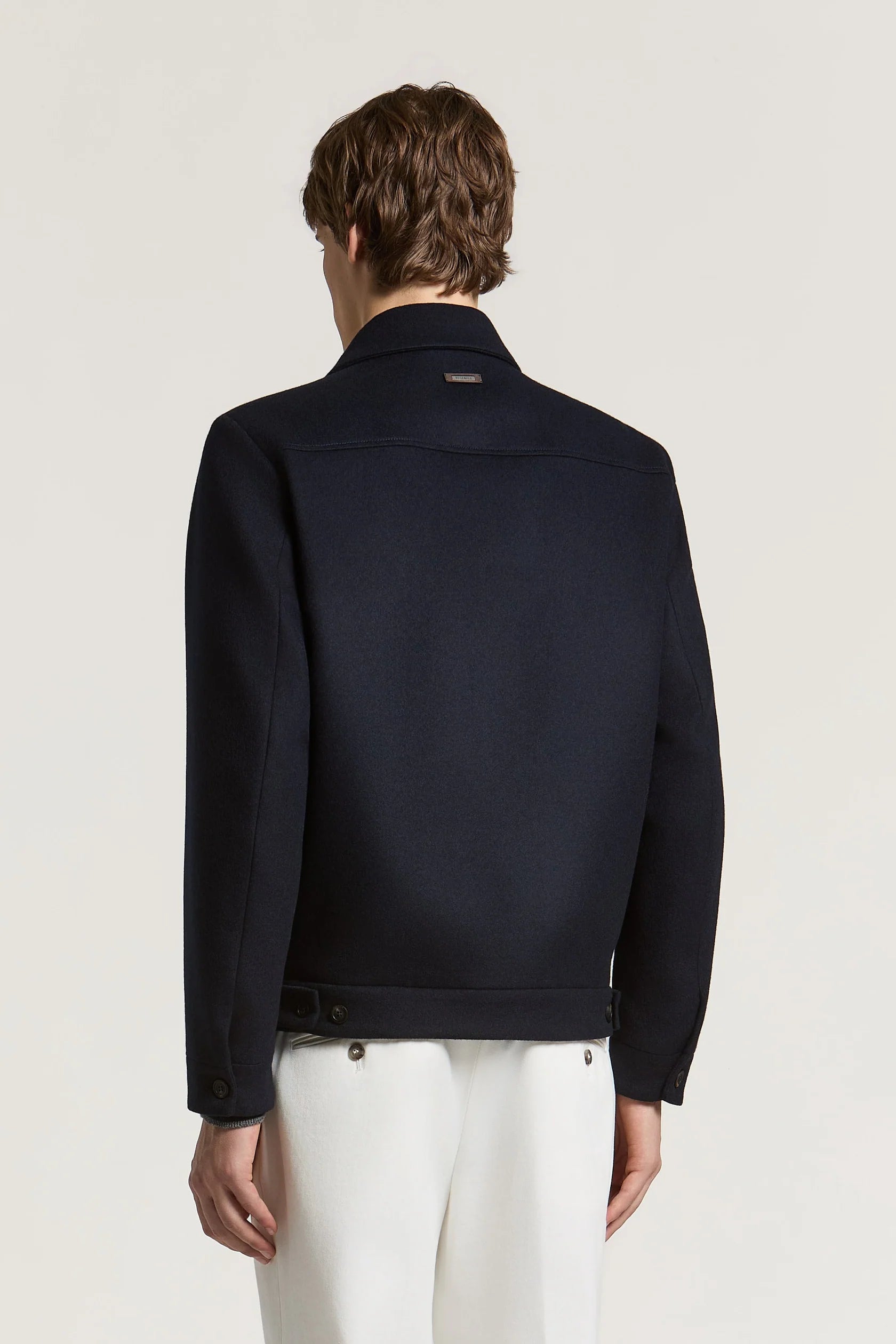 Smooth dark blue jacket with a shirt collar, back yoke, and a small gold-toned brand tag. Hem has side tabs with dark buttons