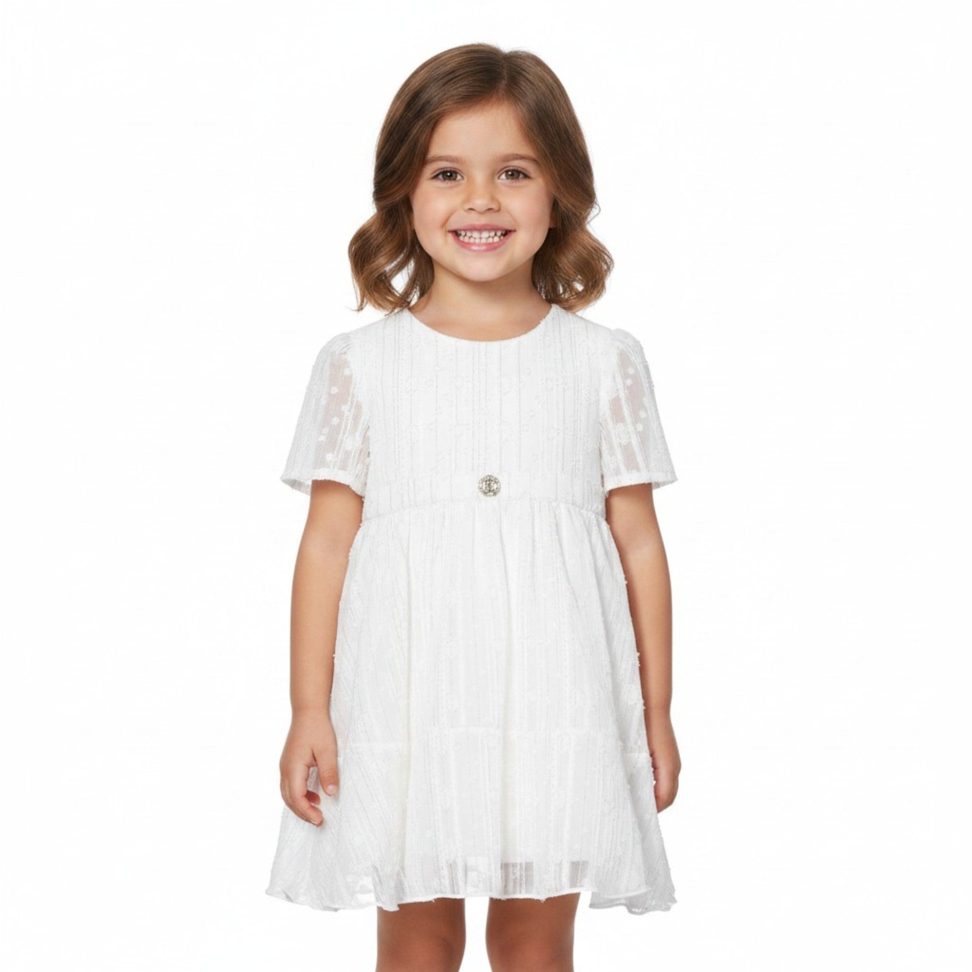 Front view of a young smiling girl with wavy brown hair modeling the knee-length white and silver Roberto Cavalli dress against a solid white background.