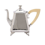 Close-up of a silver-plated teapot with a tapered body, decorative spout, and a cream-colored angular handle.