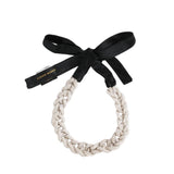 A chunky braided rope collar in a light beige/cream tone, tied with a black satin ribbon bow. A black paper brand tag with gold lettering is tucked under the ribbon, reading 'ZUNAIR HUSNAD'.