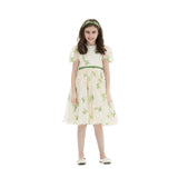 A young girl smiling in a cream tulle knee-length party dress with white floral embroidery, green waistband, and matching headband against a white background.
