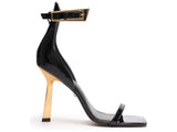 Side profile of Schutz black patent leather sandal featuring a gold stiletto heel and oversized gold ankle buckle.