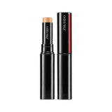 An image of a Shiseido foundation stick in a sleek, black cylindrical tube with the SHISEIDO logo in white. The open tube reveals a tan-colored cream product, and its accompanying black cap is shown beside it, featuring Shiseido's signature red vertical stripe.