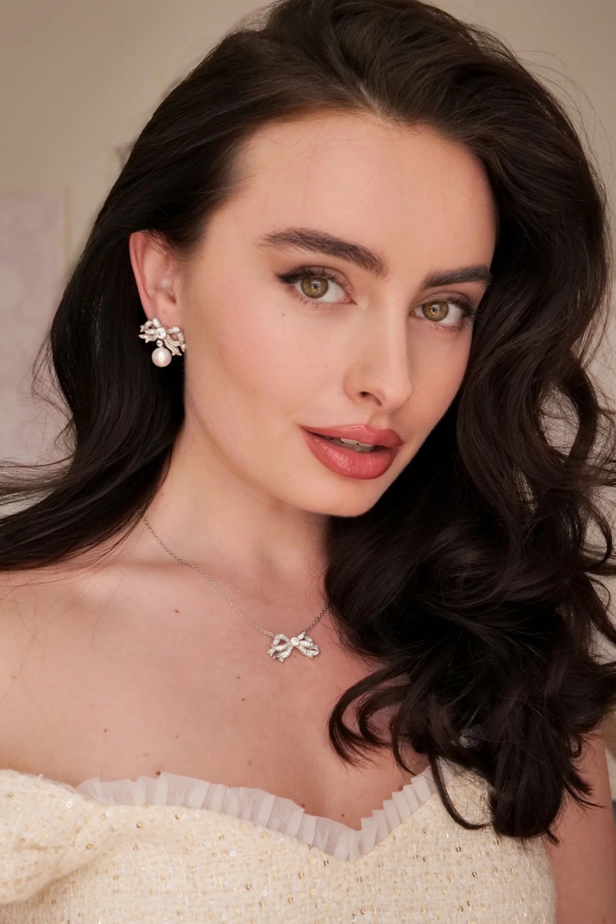 Silver-toned earring featuring a bow design encrusted with clear stones, with a white pearl dangling below.