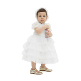 A toddler girl wearing a white tiered lace formal dress and a matching ruffled bonnet, standing against a white background.