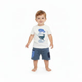 Full-length studio shot of a smiling toddler boy modeling a Stella McCartney Kids off-white t-shirt with blue whale graphics and matching slate blue jersey shorts.