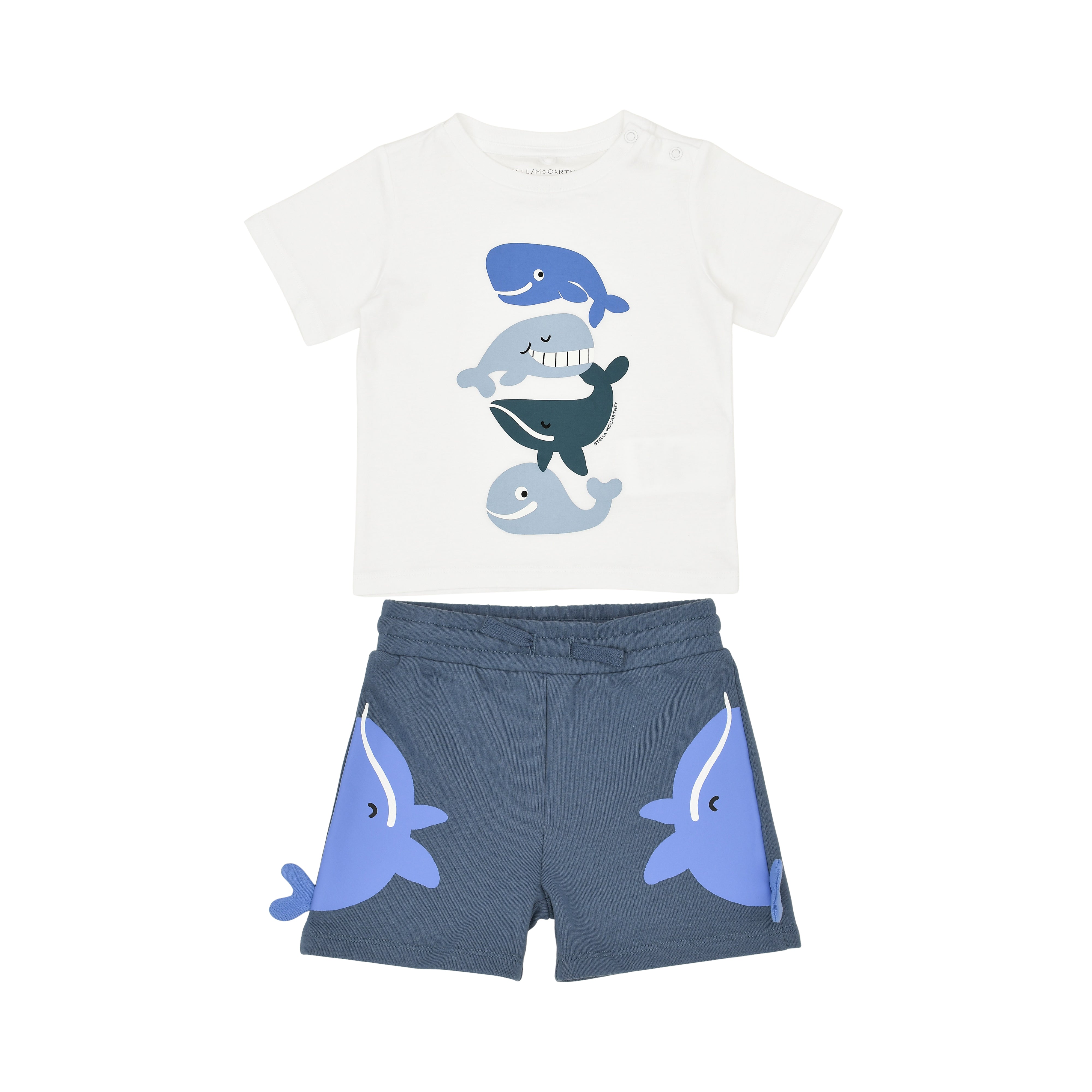 Front flat-lay view of a Stella McCartney Kids two-piece baby boy outfit consisting of a white short-sleeve t-shirt with stacked whale illustrations and blue knit shorts featuring 3D whale fin details.