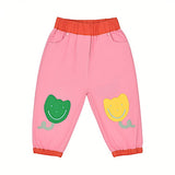 Front view of Stella McCartney Kids pink baby trousers featuring a contrasting orange elasticated waistband and ankle cuffs, front slip pockets, and a green smiling flower patch on the right leg and a yellow one on the left leg.