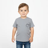 A young toddler boy standing and smiling, wearing the Stella McCartney kids' grey pocket logo T-shirt and blue jeans against a white background.
