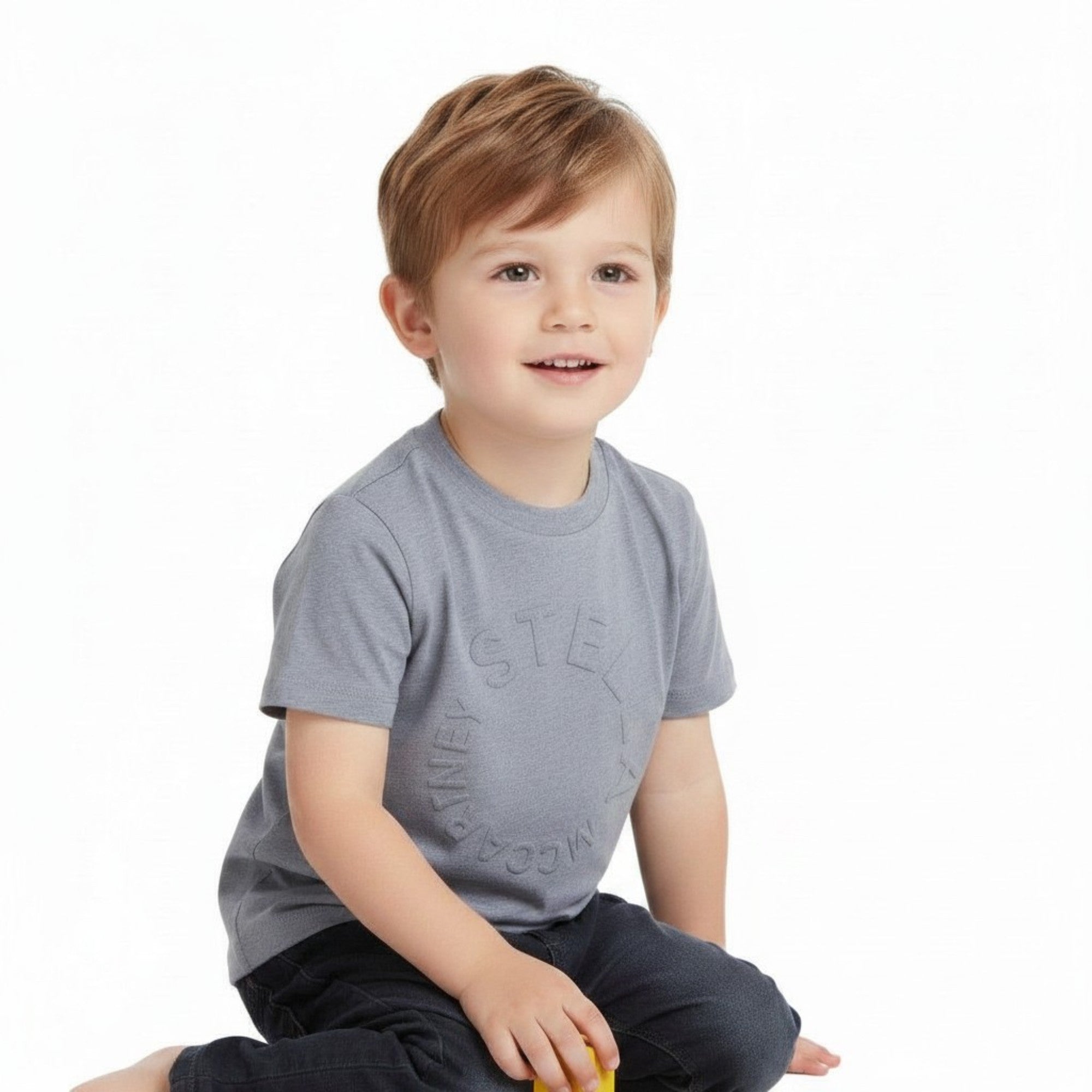 A young toddler boy sitting down, smiling and wearing the Stella McCartney kids' grey embossed logo T-shirt with dark jeans against a white background.