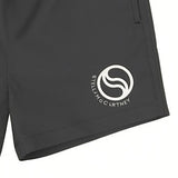 Detailed close-up of the printed white circular S-Wave graphic emblem and brand text on the lower left hem of the dark grey Stella McCartney Kids swim shorts.