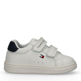 White low-cut sneaker with two velcro straps. Navy heel tab, blue 'TOMMY HILFIGER' text on top strap, and flag logo on side.