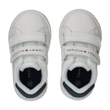 White low-cut sneakers with two velcro straps, each featuring 'TOMMY HILFIGER' text. Dark blue heel accent and light blue lin