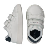 White low-cut sneakers with white velcro straps, navy blue heel accents, and a "Tommy Hilfiger" logo on the top strap of the