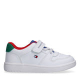 White low-cut sneaker with red trim, green heel tab, blue lining, white laces, and a Velcro strap with "TOMMY HILFIGER" text.