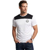 Studio shot from the waist up of a male model with short brown hair wearing the white and black Tombolini crest t-shirt, styled casually with dark blue jeans.