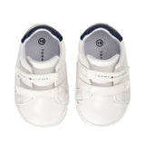 Pair of white low-cut sneakers with navy blue ankle trim and two white velcro straps. Tommy Hilfiger logo on straps.