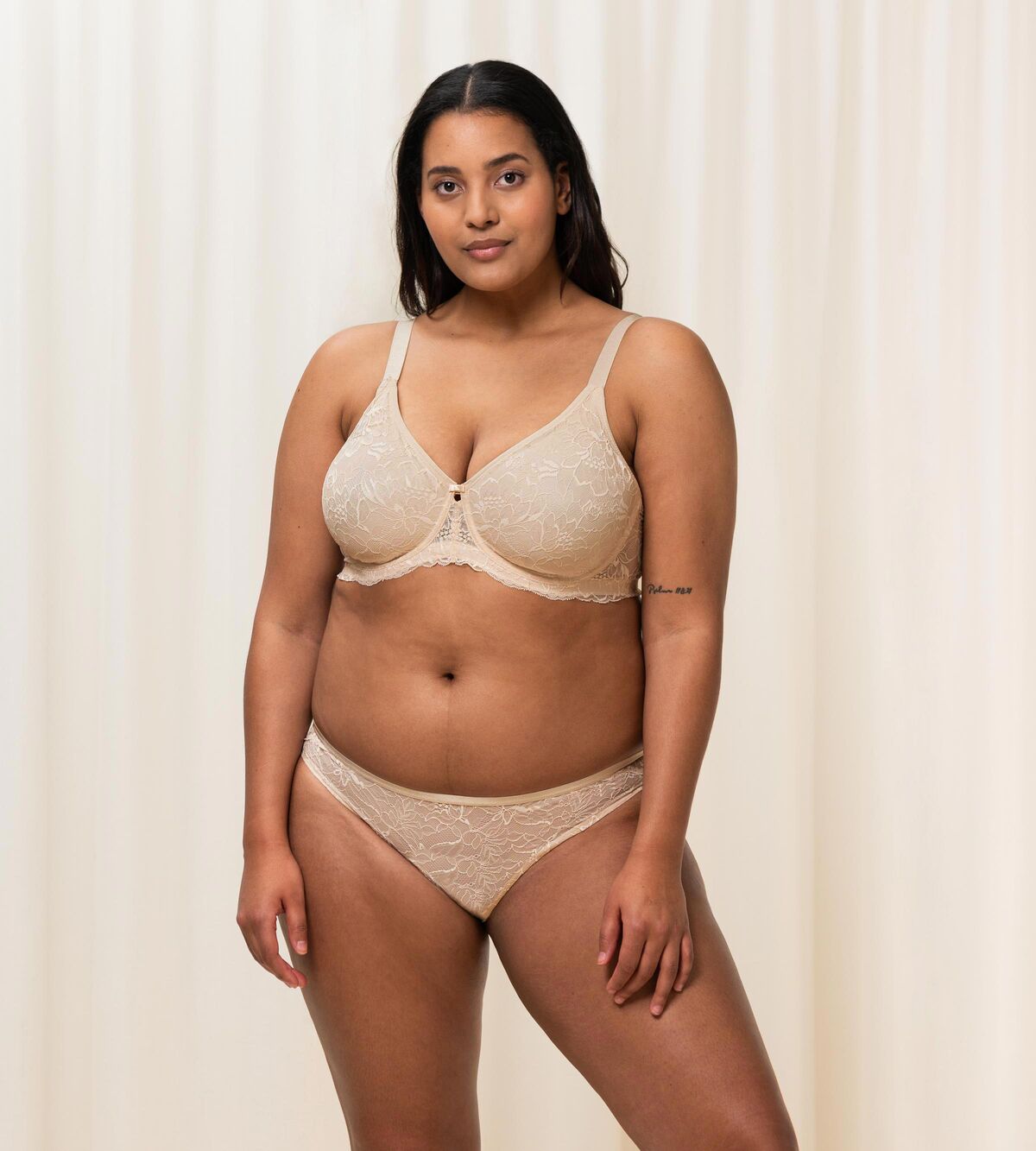 Triumph Amourette Charm Brazilian panties in light beige third model full front view, featuring semi-transparent floral lace and flat edges. Available Now at Blue Salon Qatar