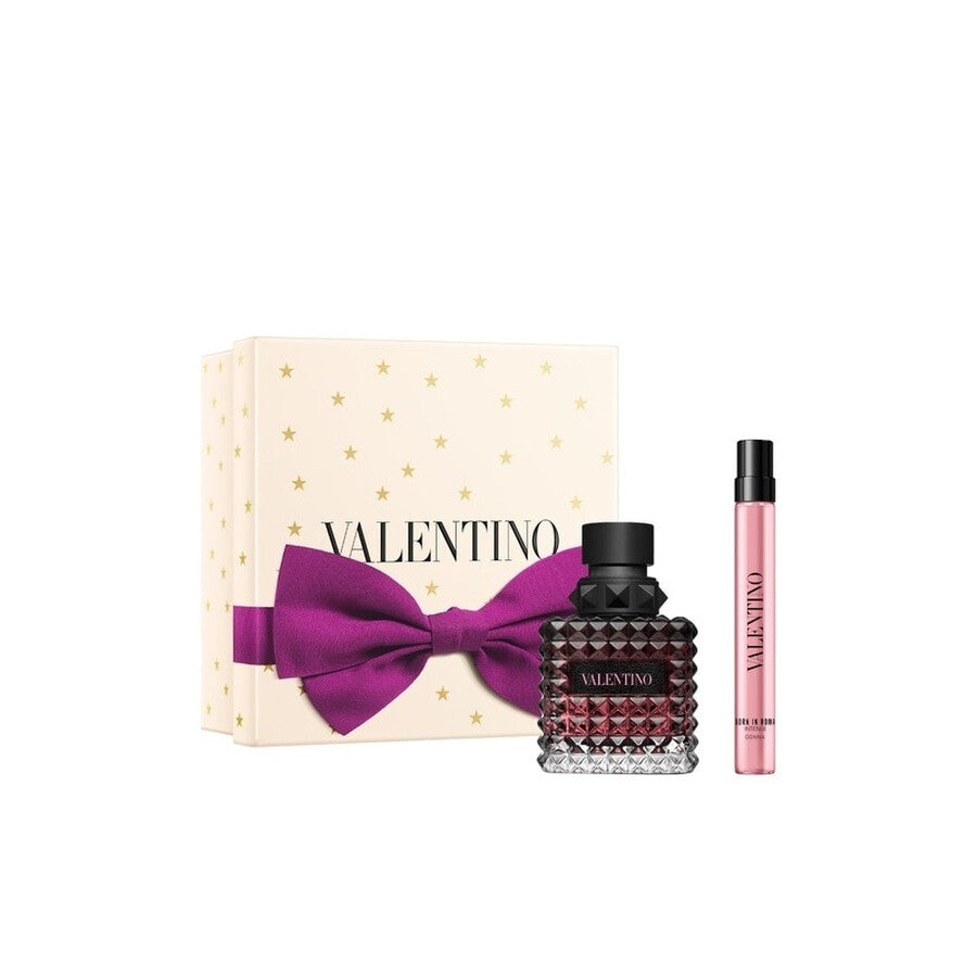 A Valentino Donna Born In Roma Intense gift set featuring a 50ml studded glass fragrance bottle with a dark burgundy gradient, a slim 15ml travel spray, and a cream-colored gift box decorated with gold stars and a large purple bow.