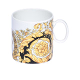 A white porcelain mug featuring a luxurious gold and black Versace Baroque pattern with ornate leaf scrolls.