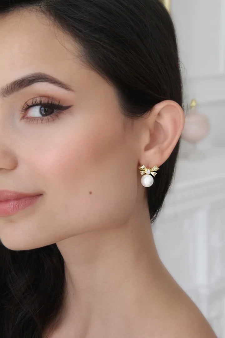 A single gold-toned earring featuring a bow studded with clear stones, from which a round white pearl dangles.