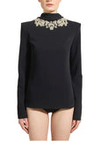 A model wearing a black long-sleeve mock-neck top with ornate silver and white beaded embellishments around the high collar, set against a plain light gray studio background.