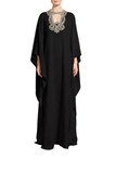A woman modeling a long black caftan dress with a wide collar embellished with silver embroidery, standing against a neutral light gray studio background.