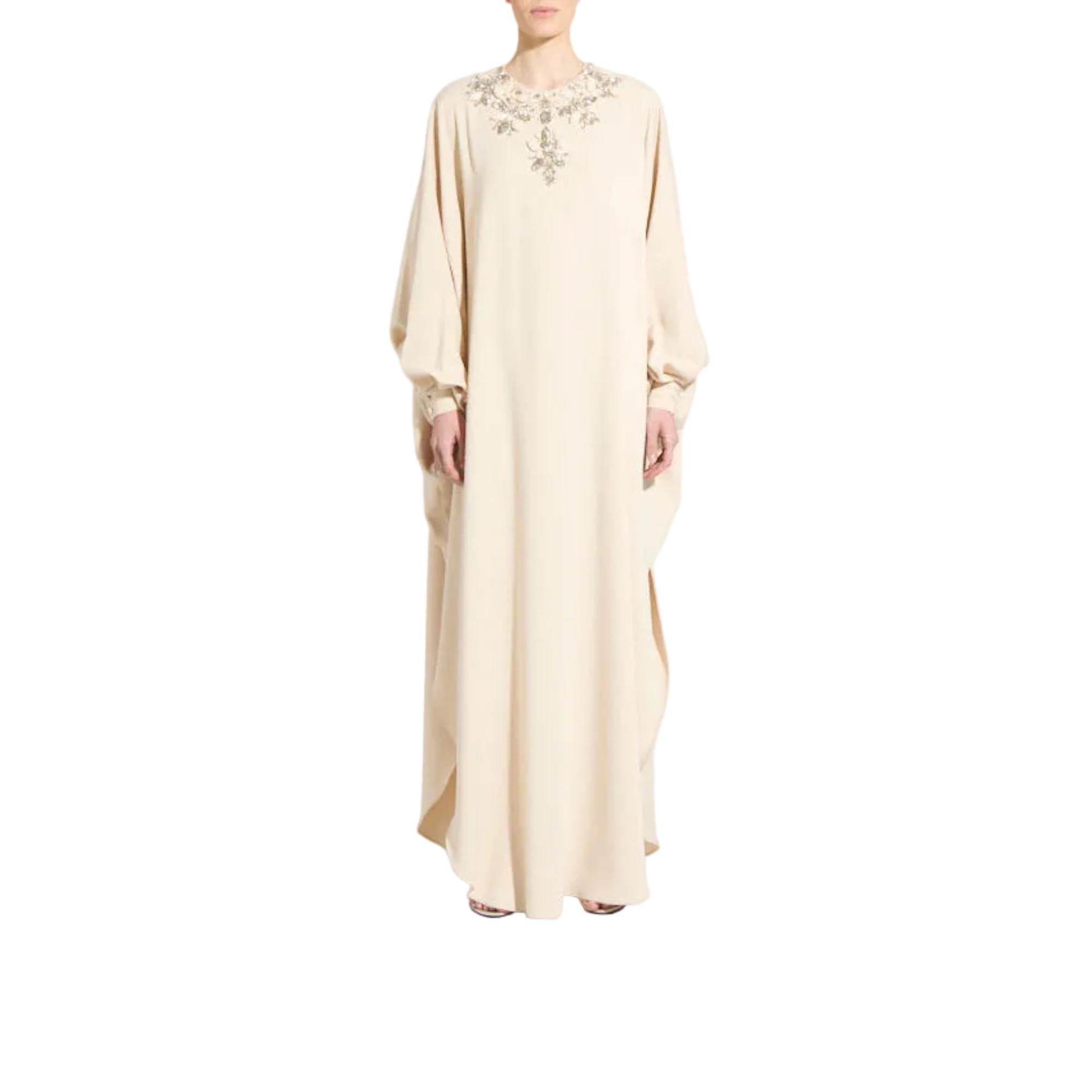 A woman modeling a long, flowing beige or cream abaya dress with wide sleeves and delicate silver-toned embroidery around the neckline, shown against a plain white background.