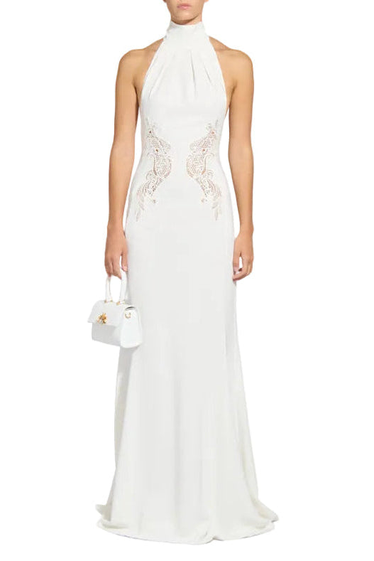 Front studio shot of a model wearing a floor-length Zuhair Murad white halter dress with a high collar and sheer, symmetrical lace side cutout inserts at the waist, holding a matching small white handbag.