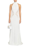 Front studio shot of a model wearing a floor-length Zuhair Murad white halter dress with a high collar and sheer, symmetrical lace side cutout inserts at the waist, holding a matching small white handbag.