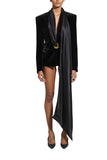 A model stands against a light gray studio background wearing a black velvet blazer with a draped satin shawl over a black mini dress, styled with black pointed-toe heels and a gold brooch.