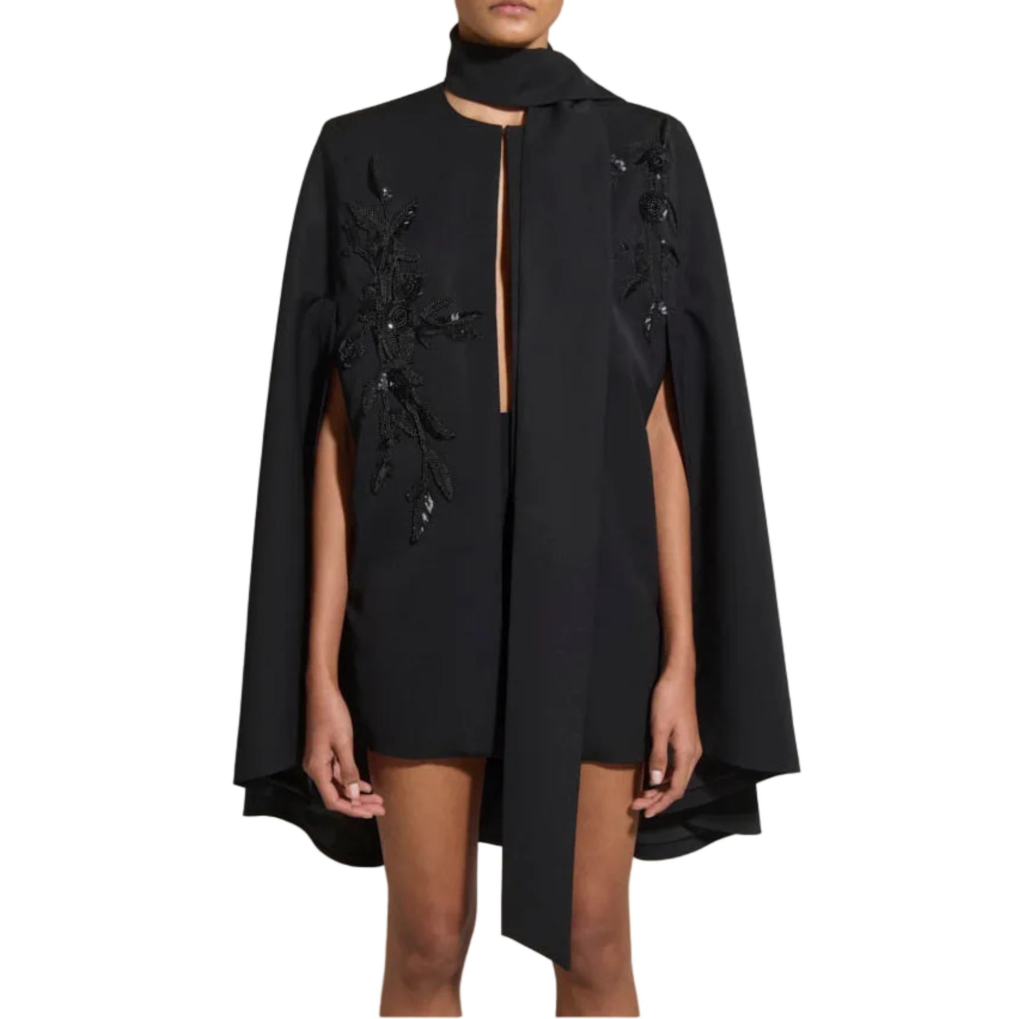 A model wearing a black high-low cape dress with a keyhole neck, side slits, and black floral bead embroidery on the front panels, set against a plain white background.
