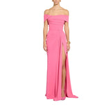 A woman modeling a bright pink off-the-shoulder floor-length gown with a fitted bodice, draped neckline, and a high front slit, shown against a plain white background.