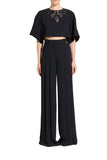 A model stands against a light gray studio background wearing a black short-sleeve crop top with a decorative embroidered neckline and matching high-waisted black wide-leg trousers.