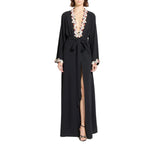 A woman modeling a long black wrap-style maxi dress with a deep V-neckline, high front slit, and white floral embroidery around the neckline and cuffs, shown against a plain white background.