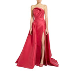 A woman modeling a strapless formal gown in bright pink-red satin. The dress features a draped bodice, a high thigh slit, and a slight train on a plain white background.